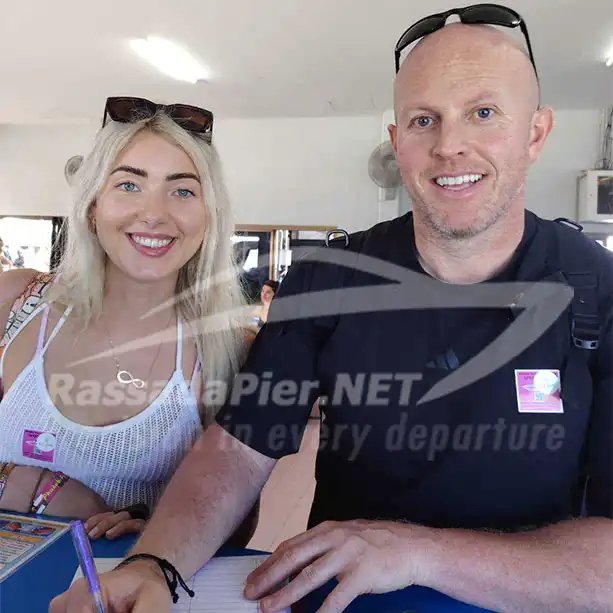 Two smiling tourists wearing pink boarding stickers while signing the passenger manifest at the Rassada Pier check-in counter